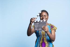 africano americano niña posando con claqueta en frente de cámara, sonriente y utilizando película pizarra en el enviar producción película industria. hembra modelo trabajando en filmografia o videografía foto