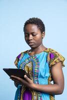 African american woman is using a modern tablet in studio, scrolling through online webpages over blue background. Girl holding device to stay connected with people on social media platforms. photo