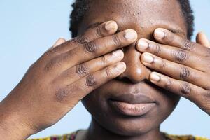 Young adult presenting three wise monkey sign in studio, covering her eyes to not see anything. African american woman recreating motivational iconic metaphor, famous emoji. photo