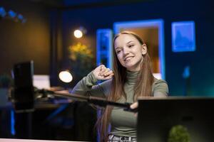 Joyful Gen Z girl records introductory video with smartphone on selfie stick, inviting viewers to subscribe to channel. Upbeat internet star uses selfie camera to film herself connecting with fans photo