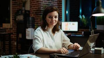 Portrait of smiling accountant researching key data for company project, doing tasks in office overnight. Happy employee sitting at desk in front of laptop, looking over accounting figures, camera A photo