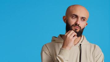Thoughtful person being puzzled about a decision, standing over blue background in studio and thinking. Pensive middle eastern man being uncertain and doubtful about something. Camera 2. photo