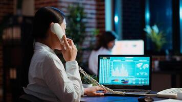 Asian employee taking landline telephone call while working with financial statistics graphs on laptop. Worker talking over the phone and solving tasks in office, doing multitasking, camera B photo