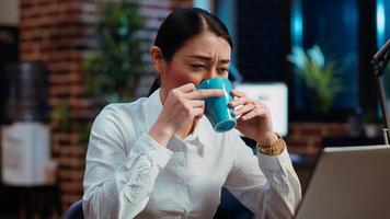 Employee tired while writing emails at computer desk, trying to freshen up by drinking coffee. Exhausted worker working overnight, typing on laptop, trying to get energy from caffeine, camera B photo