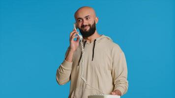 Muslim adult taking call on a landline phone in studio, using vintage office telephone with a cord. Arab guy standing over blue background and having a discussion on a remote call. Camera 1. photo