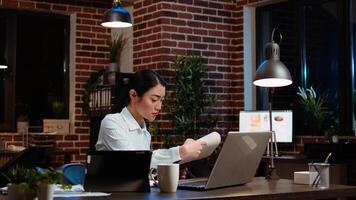 Businesswoman crosschecks data between financial charts and company internet website, doing tasks for team project in office overnight. Employee works at night, typing on laptop keyboard, camera A photo