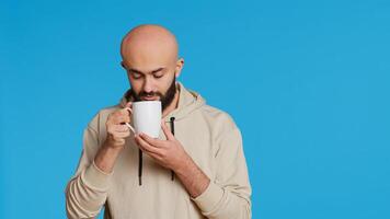 Muslim person enjoying fresh caffeine aroma in studio, drinking cup of coffee and standing over blue background. Middle eastern adult smelling beverage and taking a sip. Camera 2. Handheld shot. photo
