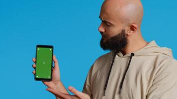 Muslim guy holding smartphone with greenscreen display, pointing at isolated chromakey template on mobile phone app. Young adult presenting blank copyspace mockup layout. Camera 2. photo
