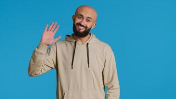 Middle eastern guy greeting people in front of camera, waving hello and smiling over blue background. Young person posing with confidence in studio, feeling confident and joyful. Camera 1. photo