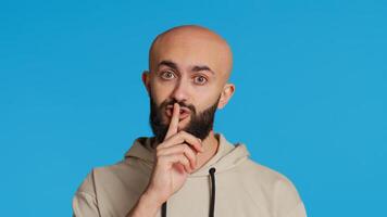 Middle eastern man doing hush silence symbol in studio, asking to keep a secret over blue background. Arab person creating mute gesture to be quiet in front of camera. Camera 2. photo