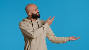 Cheerful man creating ad in front of studio camera, pointing to left or right sides over blue background. Middle eastern person showing commercial or advertisement for marketing campaign. Camera 1. photo