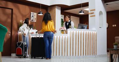 asiático turistas entrando hotel vestíbulo y utilizando Servicio campana, El sonar a llamada empleados y oferta apoyo a cheque en. viajeros solicitando asistencia desde frente escritorio personal, hospitalidad industria. foto