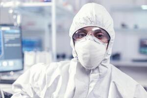 Portrait of exhausted chemist looking at camera wearing coverall in modern equipped lab. Tired woman scientist in biotechnology laboratory wearing protective suit during global epidemic. photo