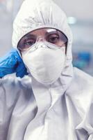 Tired laboratory doctor with overall suit looking at camera in equipped lab. Overworked woman scientist in biotechnology laboratory wearing protective suit during global epidemic. photo