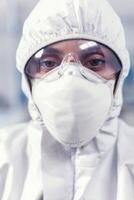 Close up of scientist woman dressed in coverall sitting at workplace in laboratory. Tired woman scientist in biotechnology laboratory wearing protective suit during global epidemic. photo