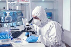 Healthcare specialist analyzing vaccine on microscope dressed in protection suit. Virolog in coverall during coronavirus outbreak conducting healthcare scientific analysis. photo