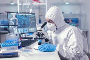 Healthcare researcher analying sample on microscope dressed in ppe suit. Virolog in coverall during coronavirus outbreak conducting healthcare scientific analysis. photo