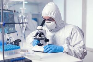 Male scientist working with microscope doing researching cure for covid19 wearing ppe suit and glasses. Virolog in coverall during coronavirus outbreak conducting healthcare scientific analysis. photo