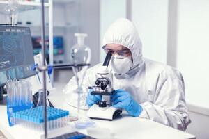 Analyzing virus in microbiology lab using microscope wearing ppe suit and glasses. Virolog in coverall during coronavirus outbreak conducting healthcare scientific analysis. photo