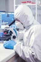 Medic in time of global pandemic working on microscope dressed in ppe suit. Virolog in coverall during coronavirus outbreak conducting healthcare scientific analysis. photo