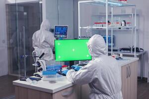 Health care reseacher dressed in ppe analyzing sample on computer with green screen in laborator. Team of microbiologists doing vaccine research writing on device with chroma key, isolated, mockup display. photo