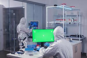 Computer with green screen in lab and medical staff dressed in ppe suit during coroanvirus. Team of microbiologists doing vaccine research writing on device with chroma key, isolated, mockup display. photo