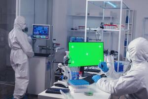 Scientist typing on keyboard works with computer with green screen dressed in ppe. Team of microbiologists doing vaccine research writing on device with chroma key, isolated, mockup display. photo