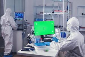 Laboratory technician in medicine lab uses computer with green screen dressed in ppe. Team of microbiologists doing vaccine research writing on device with chroma key, isolated, mockup display. photo