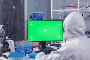 Over shoulder shot of doctor working on coroanvirus vaccine using computer with green screen dressed in ppe. Team of microbiologists doing vaccine research writing on device with chroma key, isolated, mockup display. photo