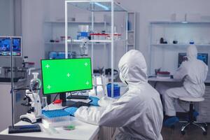Doctor in protection suit against covid19 delevoping cure using computer with green screen dressed in ppe. Team of microbiologists doing vaccine research writing on device with chroma key, isolated, mockup display. photo