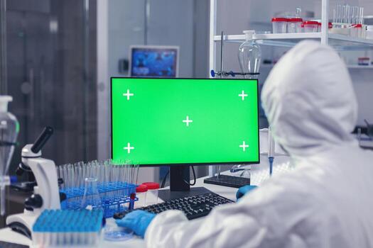 Researcher developing cure for covid19 using pc with green screen in ppe suit. Team of microbiologists doing vaccine research writing on device with chroma key, isolated, mockup display. photo