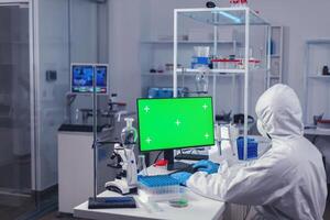 Medical researcher typing on keyboard of computer with green screen dressed in coverall. Team of microbiologists doing vaccine research writing on device with chroma key, isolated, mockup display. photo