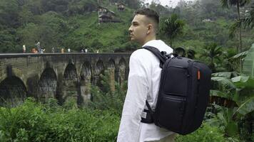 Man in jungle near bridge. Action. Man with backpack looks back at hiking trail in jungle. Handsome man on top of ancient stone bridge in jungle photo