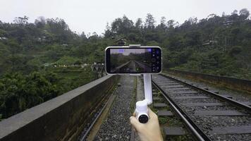 Shooting on tripod phone while traveling. Action. Shooting video on phone with stabilizer while traveling. Shooting bridge with railway in jungle on phone for blog photo