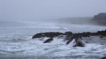 Beautiful waves break on rocks of rocky shore. Clip. Stone shore with storm waves in cloudy weather. Dramatic landscape of stone coast with beautiful splashes of waves photo