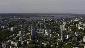 City with high-rise buildings on background of lake and green forest. Stock footage. Modern buildings in beautiful green city with lake and forest horizon. Top view of panorama of modern city in photo