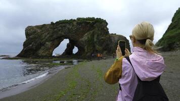 Woman on shore takes pictures of sea on phone. Clip. Woman photographs seashore with rocky arch. Group of tourists takes pictures of rocky arch on seashore photo