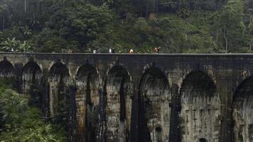 Tourists walk on stone bridge in jungle. Action. People walk on ancient stone bridge in rainforest. Beautiful landscape with people on aqueduct and green hills photo