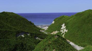 Couple walking along path on hills on background of sea. Clip. Top view of hills with trails and green grass on background of sea coast. Beautiful summer hills by sea on sunny day photo