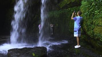 Tourists take pictures of waterfall on their phones. Clip. Tourists at water stream of rocky waterfall. Tourists at rocks with moss and waterfall in summer photo