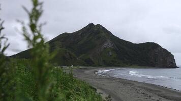 View of beautiful beach with green grass and mountain. Clip. Picturesque landscape with green grass on background of sea coast with mountain. Coast of sea with sand and rocky mountain on cloudy day photo