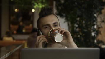 Young man is working on laptop in cafe and talking on phone. Stock footage. Young freelancer is working on laptop and answering calls. Remote work with laptop and cafe calls photo