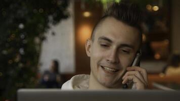 Young man is talking on phone on laptop. Stock footage. Handsome young man is talking on phone and working on laptop. Freelancer on remote with phone and laptop in cafe photo