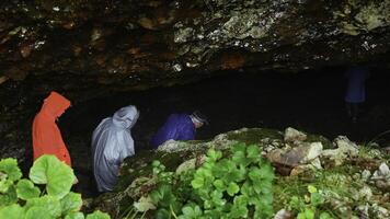 Group of tourists go to cave. Clip. Tourists enter cave in rocks on rainy day. Group of people in raincoats in rocky Mountains go to cave in summer photo