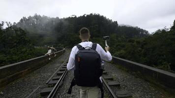 Caucasian traveler man recording video of green mountain landscape. Action. Man walking on railroad with backpack. photo
