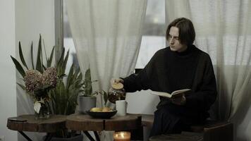 joven hombre con un taza de caliente bebida leyendo un libro a cocina, sentado en de madera silla. medios de comunicación. hombre leyendo y Bebiendo té. foto