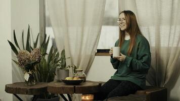 Young woman in glasses pouring tea from tea pot into a mug at home. Media. Red haired lady at kitchen drinking green tea. photo