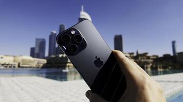 Dubai - United Arab Emirates, January 18, 2023. close up of male hand holding new Apple Iphone against water fountains in Dubai City. Action. Beautiful panorama of a modern famous city. photo