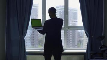 Rear view of a male student studying at home and using his laptop. Media. Man standing by the window and looking at green chroma key screen. photo