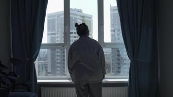 joven mujer disfrutando ver mediante el panorámico ventana en el Mañana a hogar. medios de comunicación. niña en pijama en pie y pensando por el ventana en dormitorio. foto